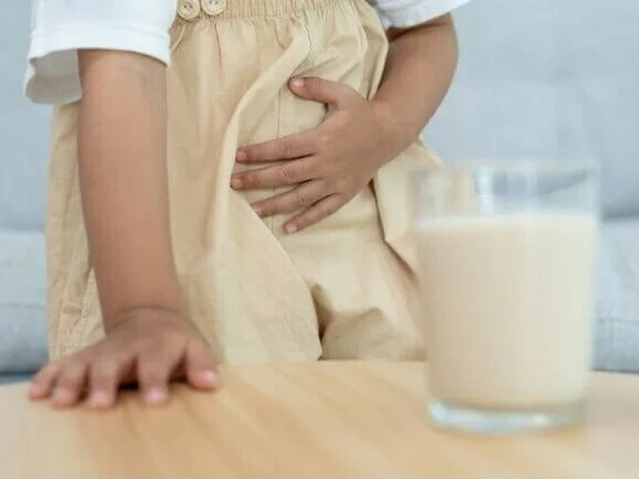Uma criança está segurando a área da barriga com uma das mãos, indicando dor ou desconforto abdominal. Um copo com um líquido que se assemelha ao leite está desfocado em primeiro plano sobre uma mesa.